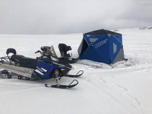 A snowmobile parked near a blue ice fishing tent on a snowy landscape, with a person preparing equipment nearby.