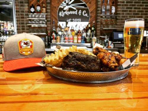 A plate of wings and fries, a beer, and a Kansas City Chiefs cap on a bar counter.