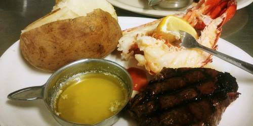 A plate featuring a baked potato, lobster tail with lemon, and a grilled steak with melted butter on the side.