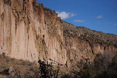 A rugged cliffside with vertical rock formations under a clear blue sky, surrounded by sparse vegetation.