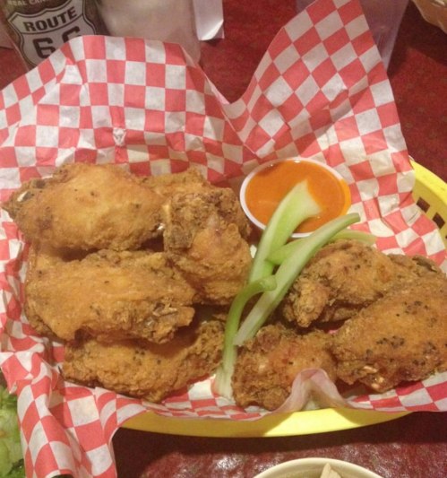 A basket of crispy fried chicken wings with celery sticks and a small cup of sauce on a checkered paper.