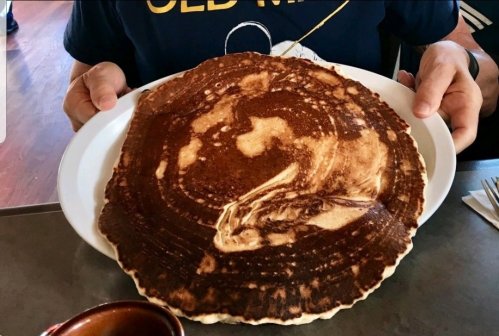 A person holds a large, round pancake on a white plate, showcasing its golden-brown surface.