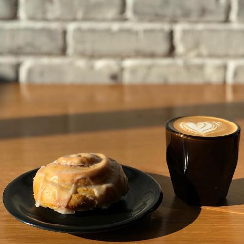 A cinnamon roll with icing on a black plate next to a heart-patterned latte in a black cup, on a wooden table.