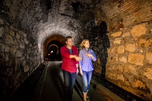 A man and woman walk together in a dimly lit stone tunnel, engaged in conversation.