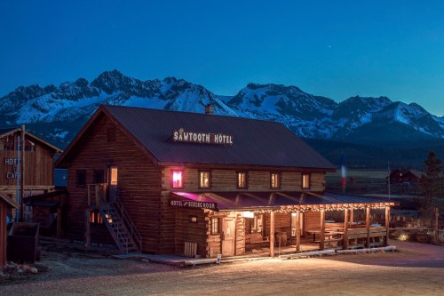 A rustic hotel with a lit sign, set against a backdrop of snow-capped mountains at dusk.