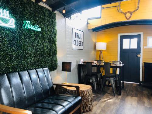 Cozy interior with a black sofa, wooden table, lamp, and greenery; features a "Trail Closed" sign and neon "Paradise" light.