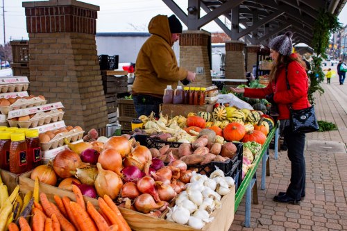 A farmer's market stall displays a variety of fresh vegetables and eggs, with a customer browsing and a vendor in a hoodie.