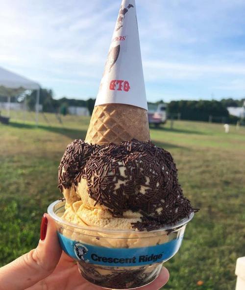 A hand holding a cone topped with chocolate sprinkles, sitting on a cup of ice cream in a sunny outdoor setting.