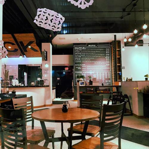 Cozy café interior with a wooden table, chairs, and a menu board featuring waffles and beverages. Warm lighting.