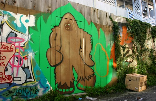 A colorful mural of a bigfoot-like creature on a wooden wall, surrounded by graffiti and greenery.