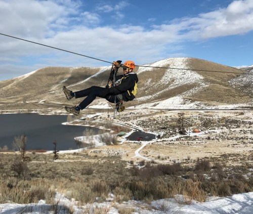 A person in a helmet zip-lining over a snowy landscape with hills and a lake in the background.