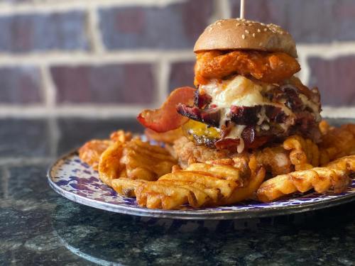A towering burger with bacon, cheese, and a sweet potato bun, served with crispy waffle fries on a decorative plate.