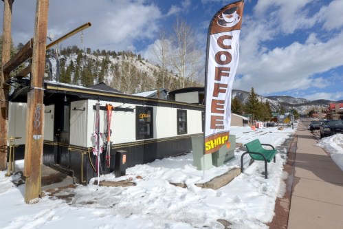 A small coffee shop with a large "COFFEE" flag, surrounded by snow and mountains on a clear day.