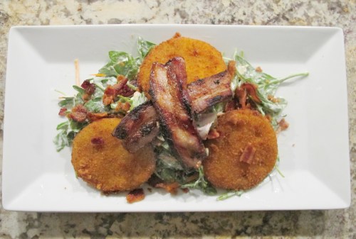 A plate of salad topped with crispy bacon, fried green tomatoes, and a creamy dressing on a textured surface.