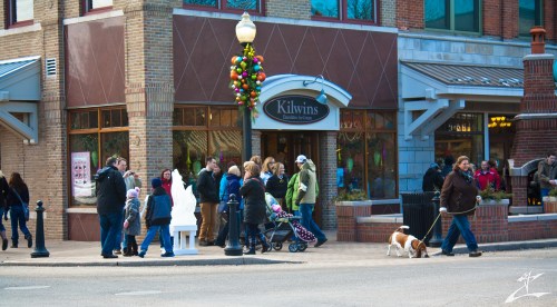 A busy street scene with a crowd outside Kilwins, decorated for the holidays, and a dog walking by.