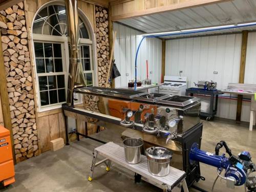 A stainless steel processing setup in a rustic kitchen, with wood stacks and various equipment in the background.