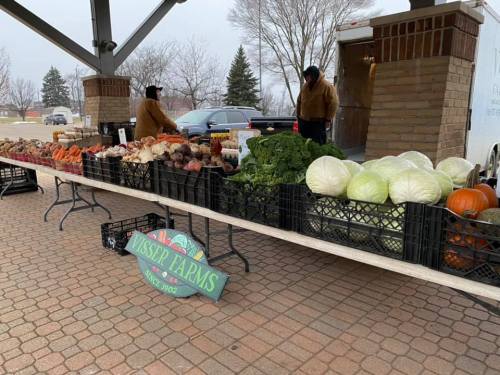 A farmers market stall displays a variety of fresh vegetables and pumpkins, with two vendors in the background.