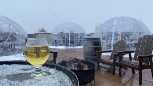 A glass of beer sits on a table near a fire pit, with igloo-like structures and snow in the background.