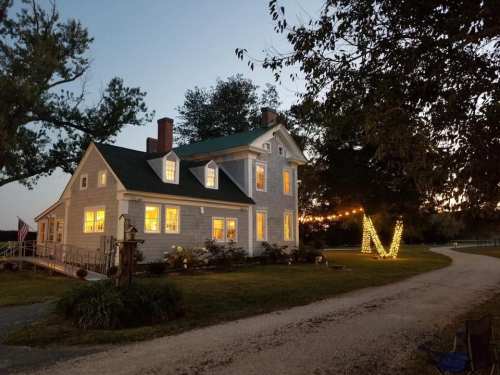 A charming house with glowing windows, surrounded by trees, and decorated with lights along a gravel path at dusk.