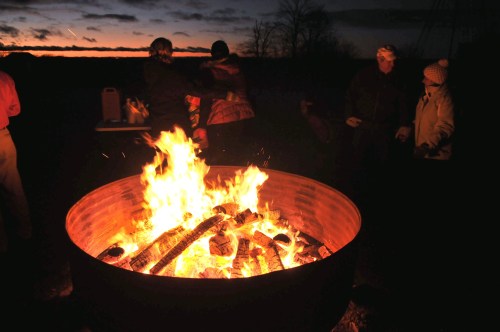 A large bonfire burns brightly at night, with people gathered around enjoying the warmth and evening sky.