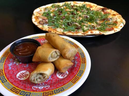 A plate of spring rolls with dipping sauce in front of a large pizza topped with herbs and vegetables.