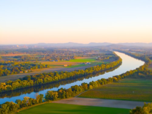 A serene river winding through lush green fields and trees, with distant hills under a soft, golden light.