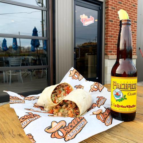A close-up of a burrito filled with meat and vegetables, next to a bottle of Pacifico beer, on a branded paper.