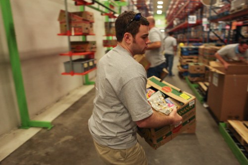 A person in a gray shirt carries a box in a warehouse filled with shelves and other workers in the background.
