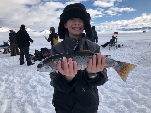 A smiling boy in winter gear holds a large fish while standing on snow, with people and mountains in the background.