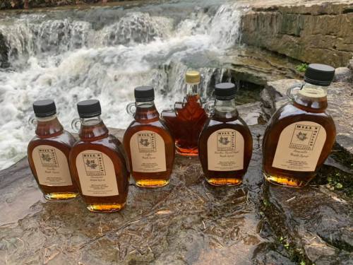 Bottles of maple syrup arranged by a waterfall, showcasing various flavors and sizes against a natural backdrop.