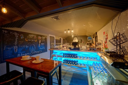 Cozy coffee shop interior with a wooden table, illuminated counter, chalkboard menu, and shelves of drinks.