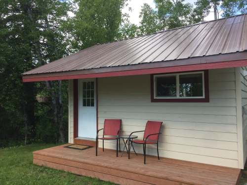 A small cabin with a metal roof, featuring a front porch and two red chairs, surrounded by greenery.