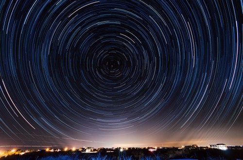 A starry night sky with circular star trails above a dark landscape, illuminated by distant lights.