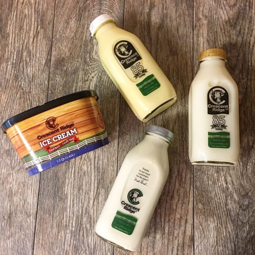 Four containers of Crescent Ridge ice cream and milk on a wooden surface, featuring a tub and three glass bottles.