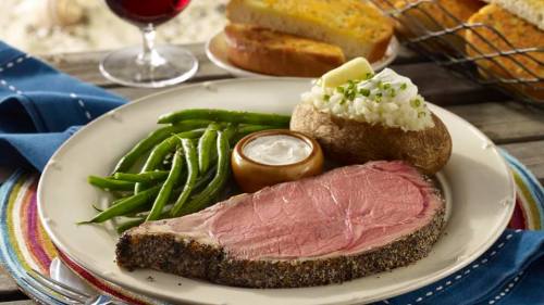 A plate featuring a slice of prime rib, green beans, a baked potato with sour cream, and toasted bread.