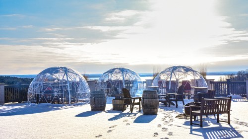 Three transparent igloos on a snowy deck overlooking a lake, with wooden barrels and chairs in the foreground.