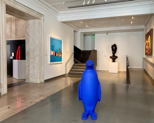 A blue penguin sculpture stands in an art gallery, surrounded by various artworks and sculptures.