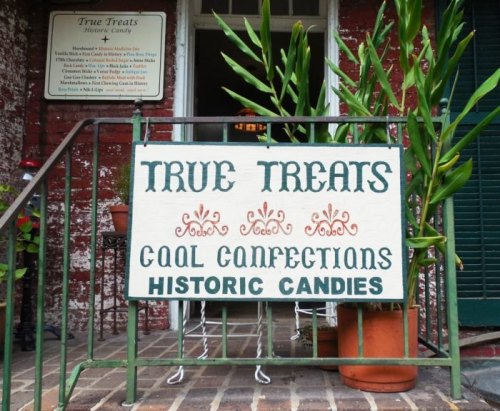 Sign for "True Treats" featuring "Gourmet Confections" and "Historic Candies," surrounded by potted plants.