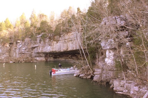 A boat near a rocky shoreline with trees, under a cliff, reflecting in calm water.