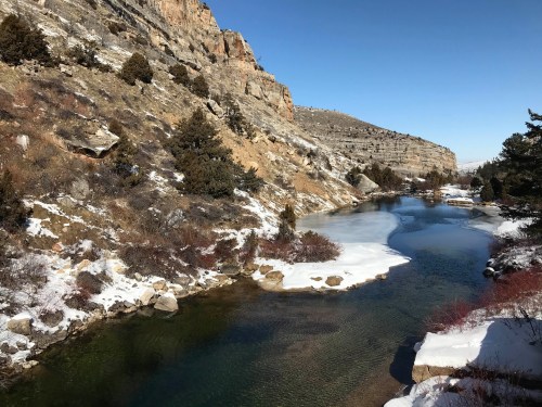 A serene river flows through a snowy landscape, flanked by rocky cliffs and sparse trees under a clear blue sky.