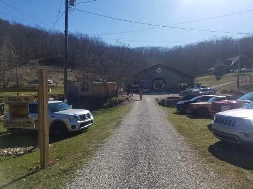 A gravel driveway leads to a rustic building with parked cars on either side, surrounded by trees and hills.