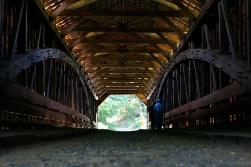 A person stands inside a covered bridge, with wooden arches and beams illuminated by natural light from the entrance.
