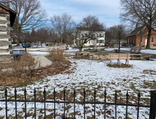 A snowy park scene with benches, trees, and historic buildings under a clear sky. Fenced area in the foreground.