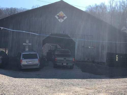 A rustic wooden barn with a geometric design on the front, parked vehicles in the foreground, and trees in the background.