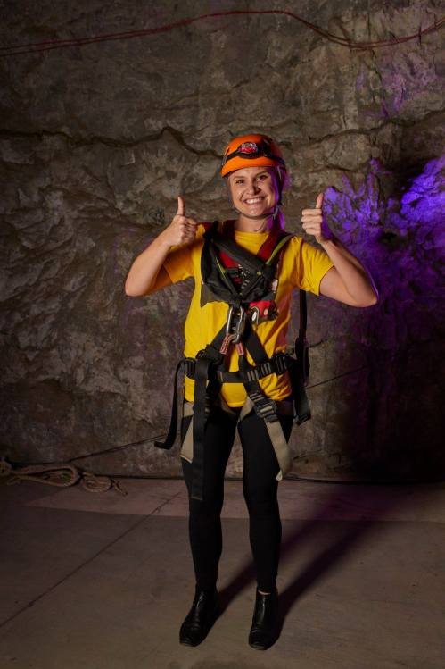 A smiling woman in a yellow shirt and helmet gives two thumbs up, wearing a harness in a rocky cave setting.