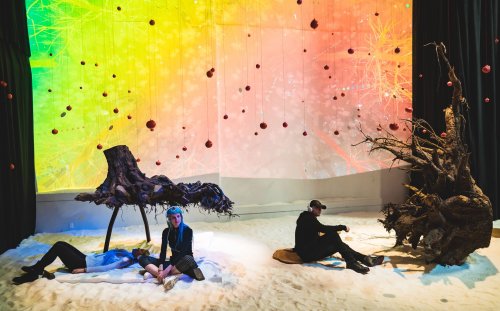 Art installation featuring three figures on a sandy floor, surrounded by colorful projections and hanging decorations.