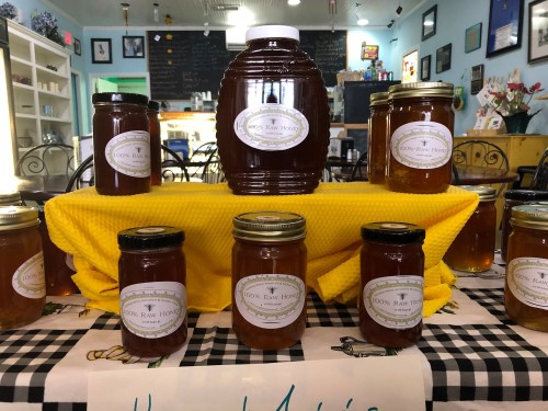 A display of various jars of 100% raw honey on a yellow cloth, set in a cozy shop with a checkered tablecloth.