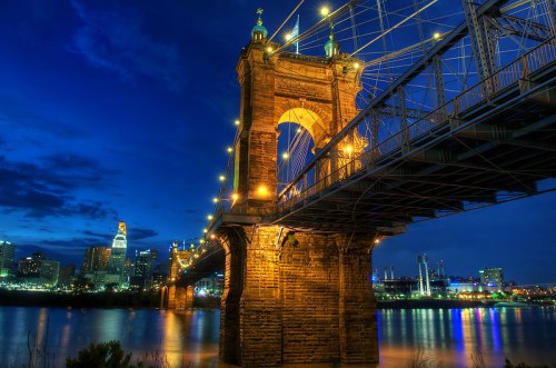 A nighttime view of a historic bridge illuminated against a city skyline, reflecting in the water below.