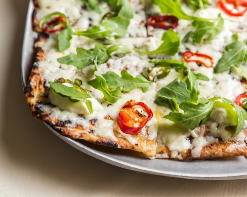A close-up of a pizza topped with melted cheese, fresh arugula, and sliced red chili peppers on a metal tray.