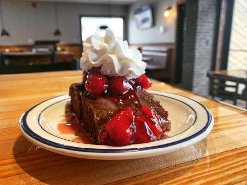 A slice of chocolate cake topped with whipped cream and cherries on a white plate, set on a wooden table.